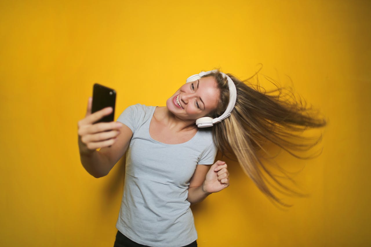 Cheerful woman wearing headphones, enjoying music and taking a selfie against a vibrant yellow background.